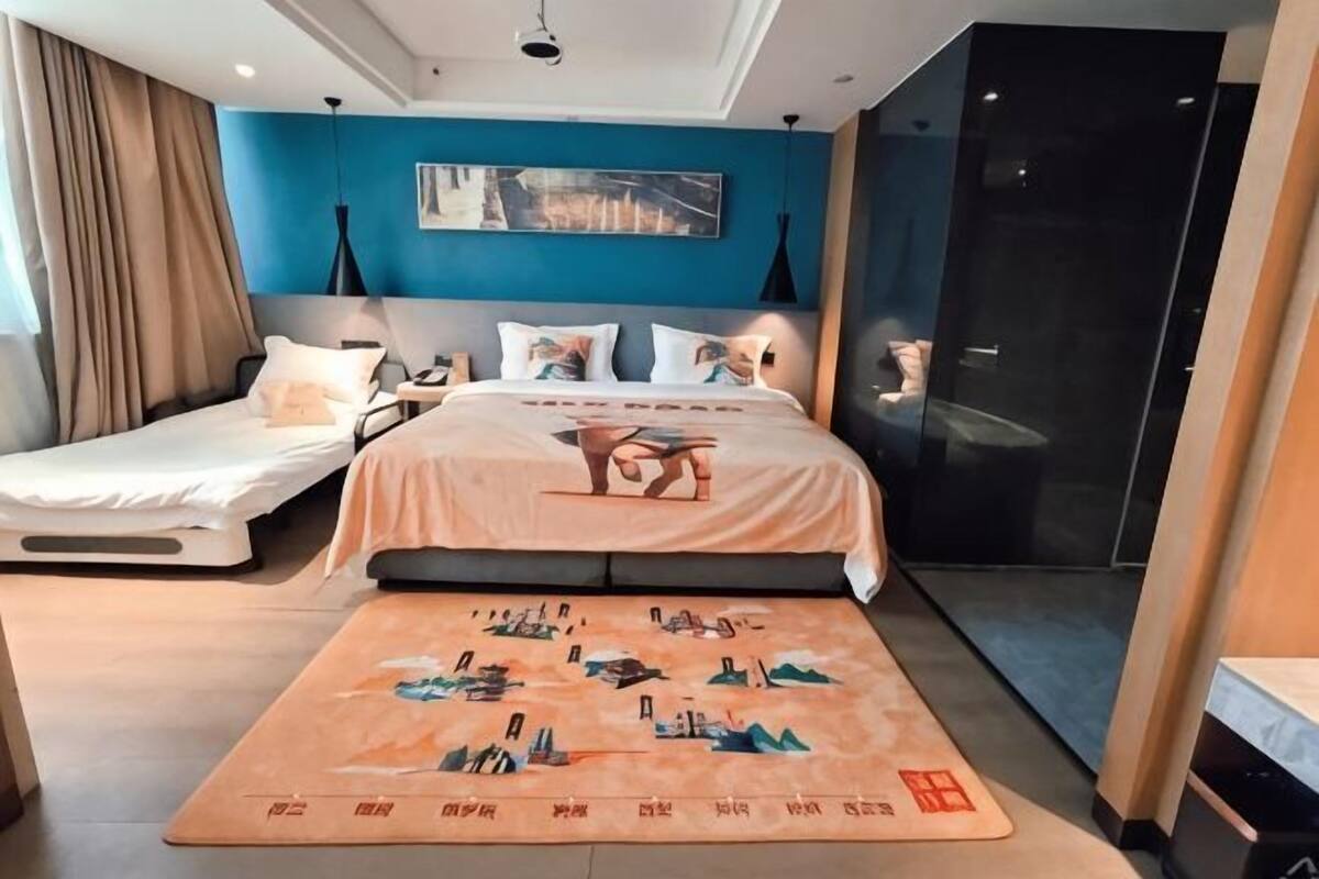 Quarto com cama, sofá-cama, mesa de cabeceira, luminária na parede e uma obra de arte com vista para a cidade.