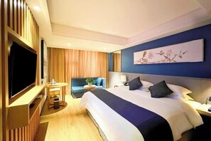 A hotel room with a large bed, a flat-screen TV, a desk with a chair, a sofa, and a painting on the wall.