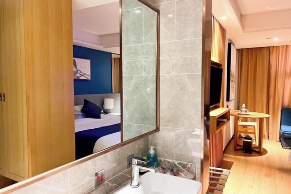 Una habitación de hotel moderna con un espejo grande, una cama con ropa de cama azul, un baño con lavabo y espejo, y una pequeña zona de estar con una mesa y una silla.