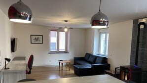 A modern living room with a black leather sofa, a wooden coffee table, and a large marble countertop. There are two pendant lights, a fireplace, and a window with blinds. - City center apartment (La Bresse)