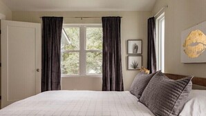 A bedroom with a large bed, dark curtains, a window with a view of greenery, and a framed artwork on the wall. - The Studio at Cleveland Park 2 bedroom 2 bathroom Near Lower Broadway (Nashville)