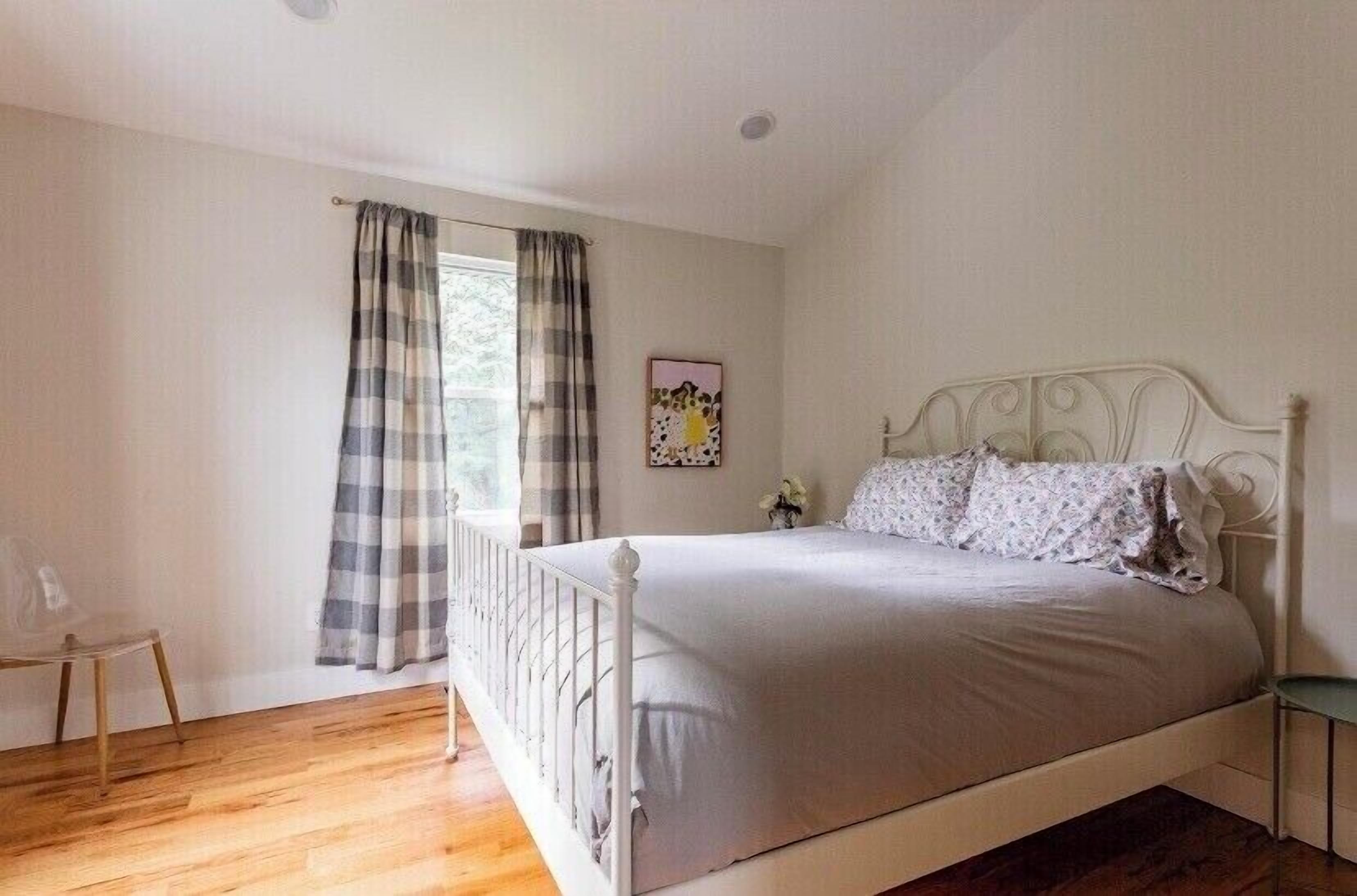 A bedroom with a white bed, a wooden floor, a window with curtains, and a small bedside table.