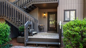 Un escalier en bois mène à une porte portant une plaque avec un numéro, et on y trouve une petite table et un banc devant l’entrée d’un bâtiment.