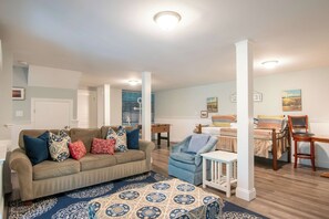 A living room with a sofa, armchair, and ottoman, a small table, and a bed with a striped bedspread.