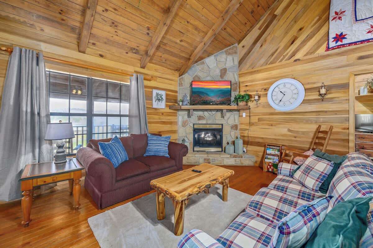 A cozy cabin interior with a stone fireplace, wooden ceiling, and plaid furniture.