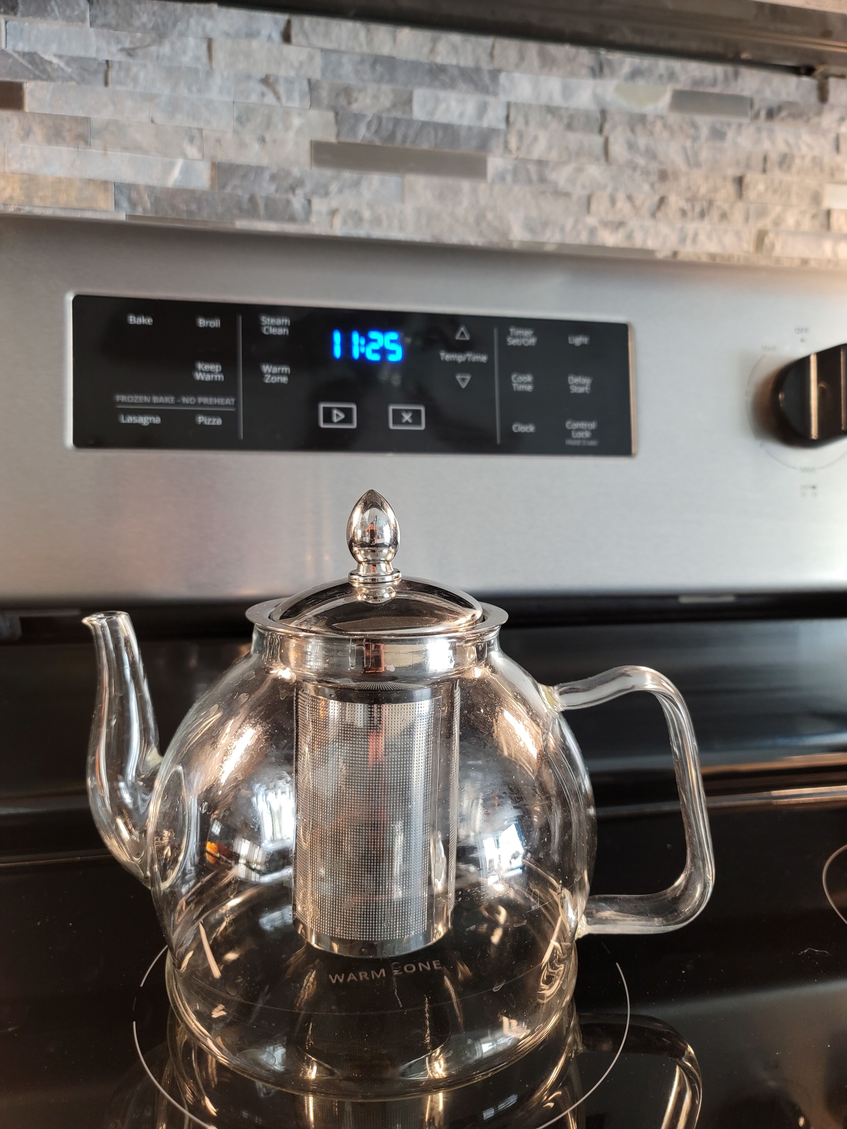 A kitchen with a digital clock displaying 11:25, a stainless steel kettle, and a stone backsplash.