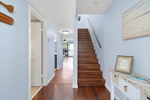 A hallway with a wooden staircase, a door leading to a room, and a framed welcome sign on a console table.