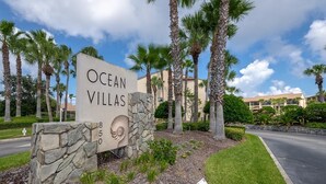 Une enseigne pour Ocean Villas, avec une base en pierre et un motif en forme de coquillage, entourée de palmiers et d’une pelouse bien entretenue.