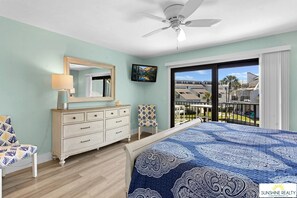 Un dormitorio con cama, cómoda, espejo, televisor y balcón con vista a edificios.