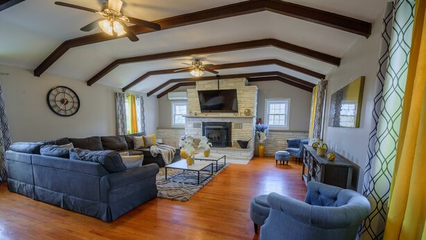 A spacious living room with a fireplace, wooden beams, a ceiling fan, and a large TV. - "Modern 6BR Pocono Lake View Home Near Attractions W Balcony” (Covington Township)
