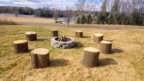 A circular fire pit surrounded by eight tree stump stools in a grassy field with a lake and trees in the background. - "Modern 6BR Pocono Lake View Home Near Attractions W Balcony” (Covington Township)