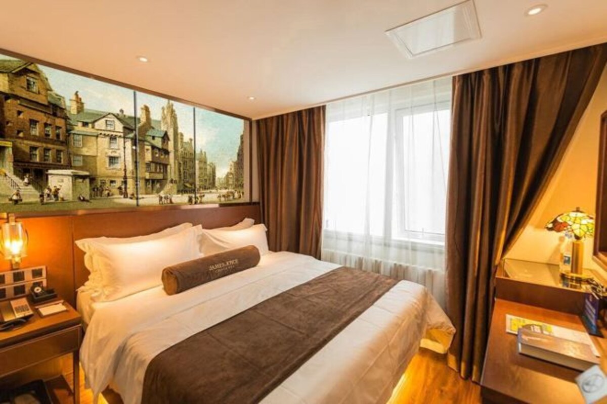 A hotel room with a large bed, a nightstand with a lamp, a desk with a computer, and a painting of a cityscape on the wall.