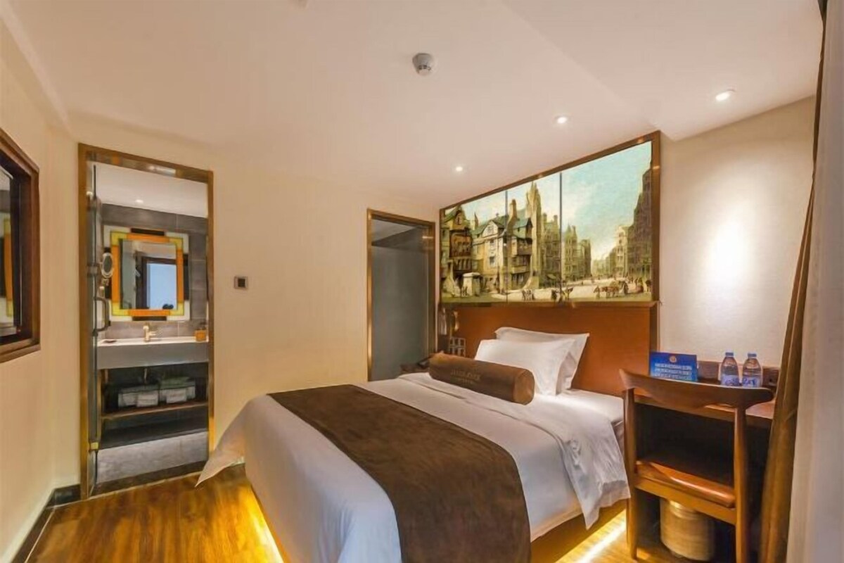 Une chambre d’hôtel avec un lit, un tableau représentant un paysage urbain, un miroir et une petite table sur laquelle se trouvent des bouteilles d’eau.