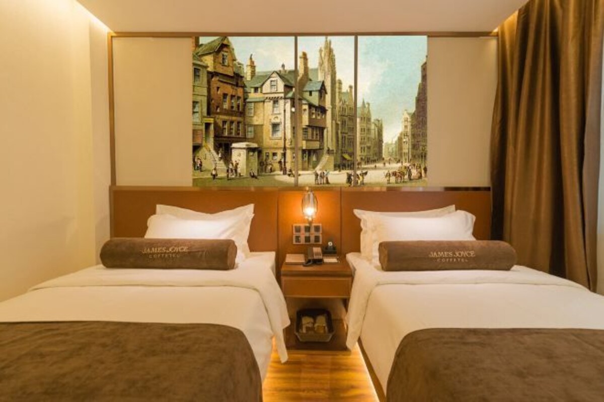 A hotel room with two beds, a nightstand with a lamp, and a large painting of a city street on the wall.