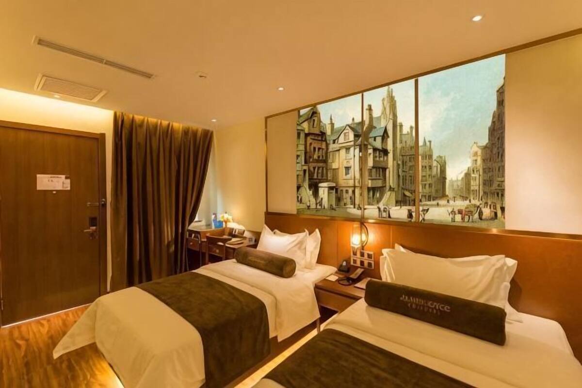 A hotel room with two beds, a large window with a cityscape view, a door, and a nightstand with a lamp.