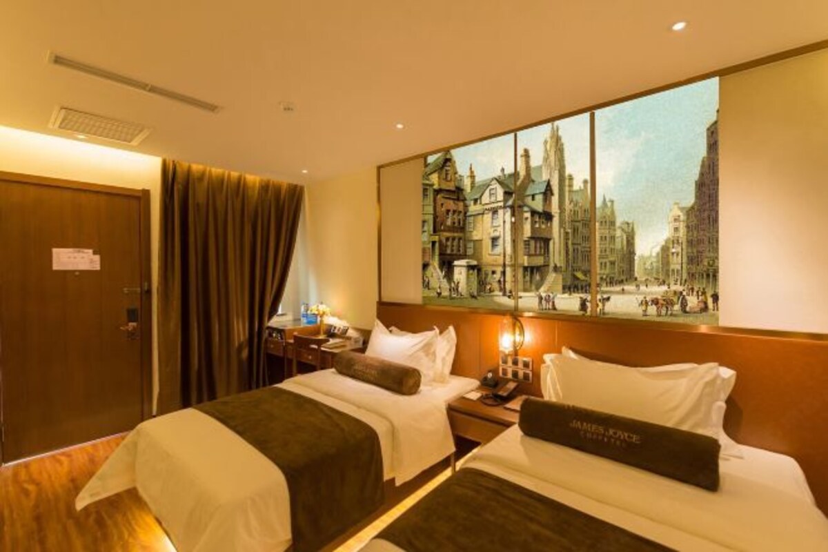 A hotel room with two beds, a painting of a cityscape, a wooden door, and a nightstand with a lamp.