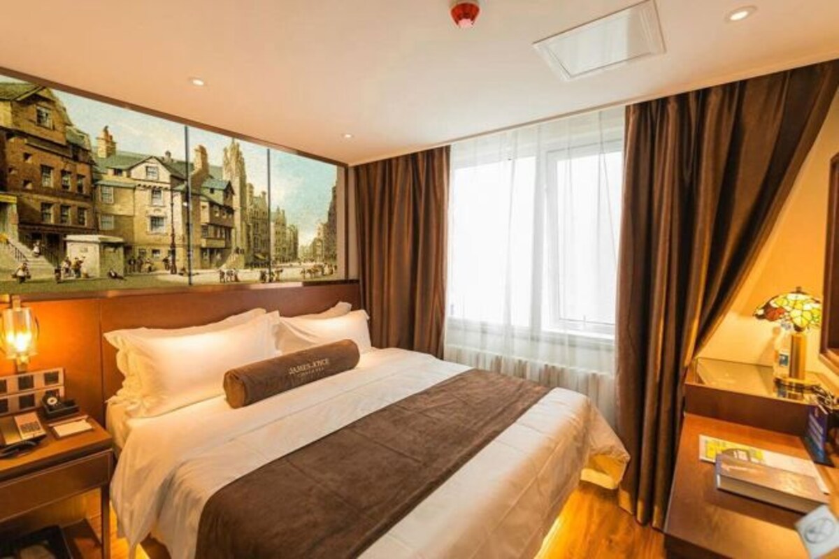 A hotel room with a large bed, a nightstand with a lamp, a bedside table with a vase, and a painting of a cityscape on the wall.