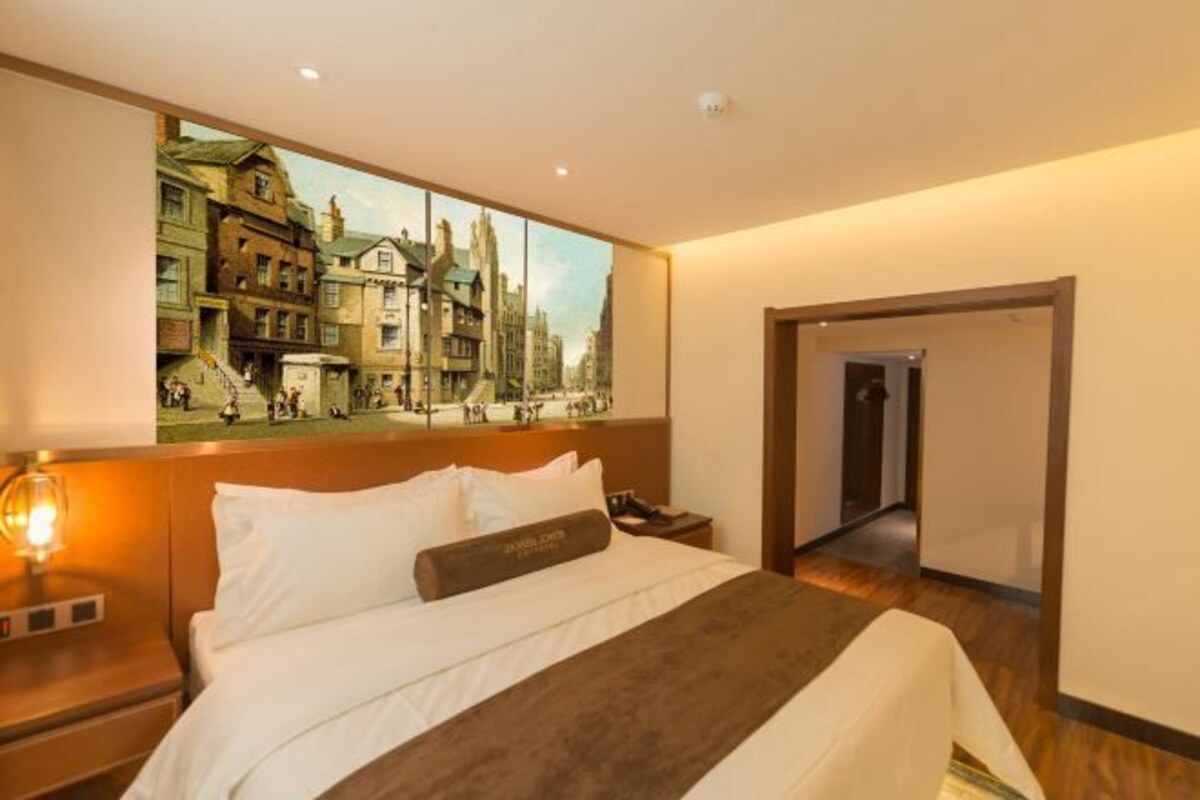 A hotel room with a large bed, a nightstand, a wall-mounted painting of a street scene, and a door leading to another room.