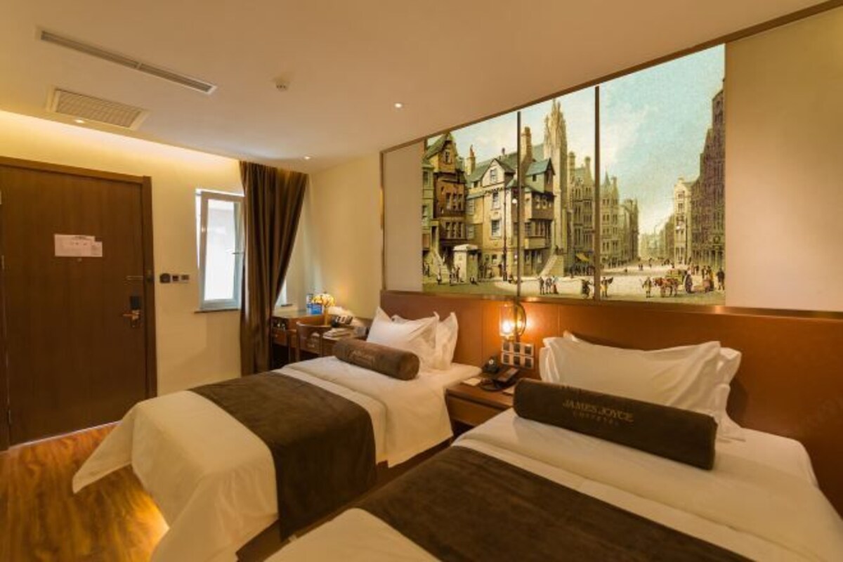 A hotel room with two beds, a painting of a cityscape, a nightstand with a lamp, and a sign on the door.
