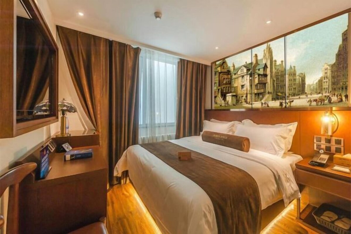 Une chambre d’hôtel avec un grand lit, une table de chevet avec une lampe, un tableau représentant un paysage urbain accroché au mur et une fenêtre avec des rideaux.