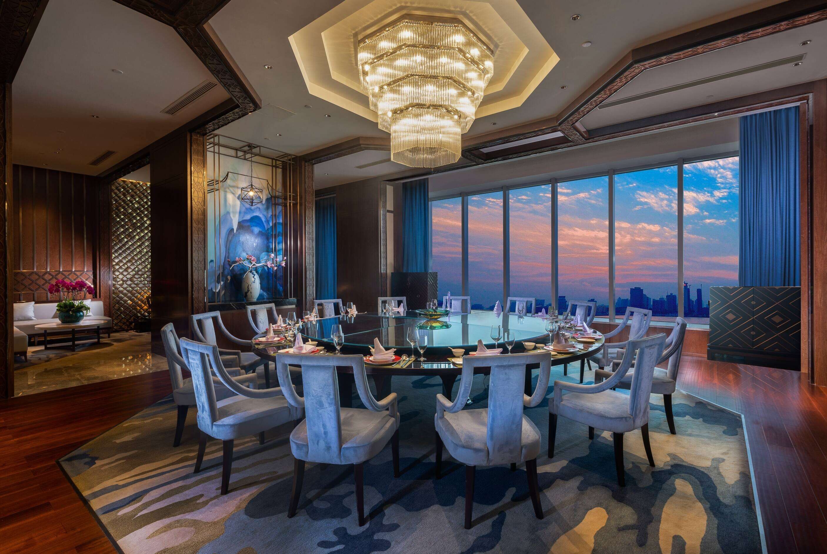 Un comedor amplio con una gran mesa redonda dispuesta para una comida, rodeado de sillas modernas y un ventanal con vista al horizonte de la ciudad.