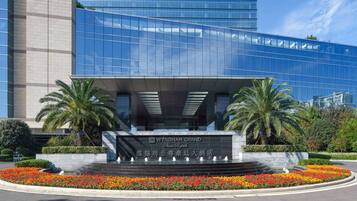 Un edificio moderno con fachada de vidrio, una entrada central con fuente de agua y palmeras.