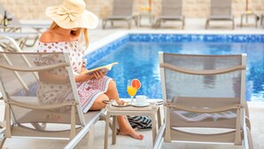 Uma mulher lendo um livro numa cadeira à beira da piscina, com uma bebida e um prato de comida.