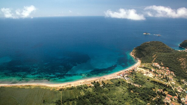 Vista aérea de uma cidade costeira com uma praia, águas azuis cristalinas e uma paisagem montanhosa.