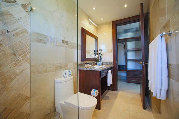 A modern bathroom with a glass shower enclosure, a marble-tiled floor, a wooden vanity with a mirror, and a white toilet. - Owner-Rented Contemporary All-Inclusive Tropical Villa (Puerto Plata)