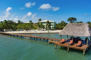 A wooden pier leading to a thatched-roof structure with lounge chairs, a beach with palm trees, and a multi-story building in the background.