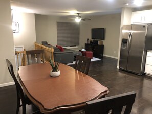 Un coin repas avec une table et des chaises en bois, une plante en pot, un canapé gris et un téléviseur fixé au mur.