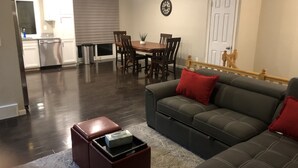 A modern living room with a dark wood floor, a grey sofa with red cushions, a dining table with chairs, a kitchen area with a refrigerator, and a clock on the wall. - MileHi Vacation Home (near DIA, pet friendly, has a baby crib) (Aurora)
