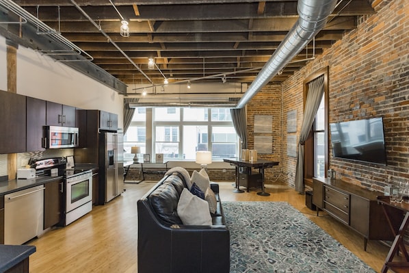 Exposed brick walls and high ceilings create an industrial atmosphere, featuring a kitchen with stainless steel appliances and an oven, a comfortable sofa, and a large flat-screen TV.