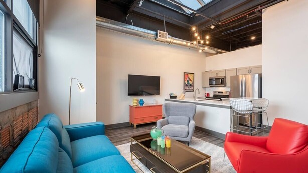 A modern living room with a blue sofa, a red chair, and a coffee table. There is a kitchen area with a microwave and a refrigerator. - Fresh, Sleek, and Contemporary Executive Apartment (Rochester)