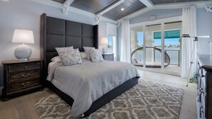 Une chambre avec un grand lit, une tête de lit sombre et un ventilateur de plafond. Il y a un balcon avec vue sur la ville.