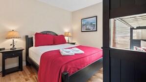 Une chambre à coucher avec un lit, deux tables de chevet avec des lampes, une couvre-lit rouge et une fenêtre avec des stores.
