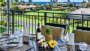 Mesa preparada para comer con botella de vino, piña, flores y velas en un balcón con vistas a un jardín exuberante y al océano.