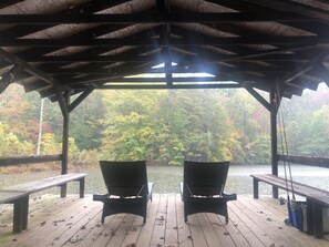 A covered wooden deck with two chairs facing a lake surrounded by trees.