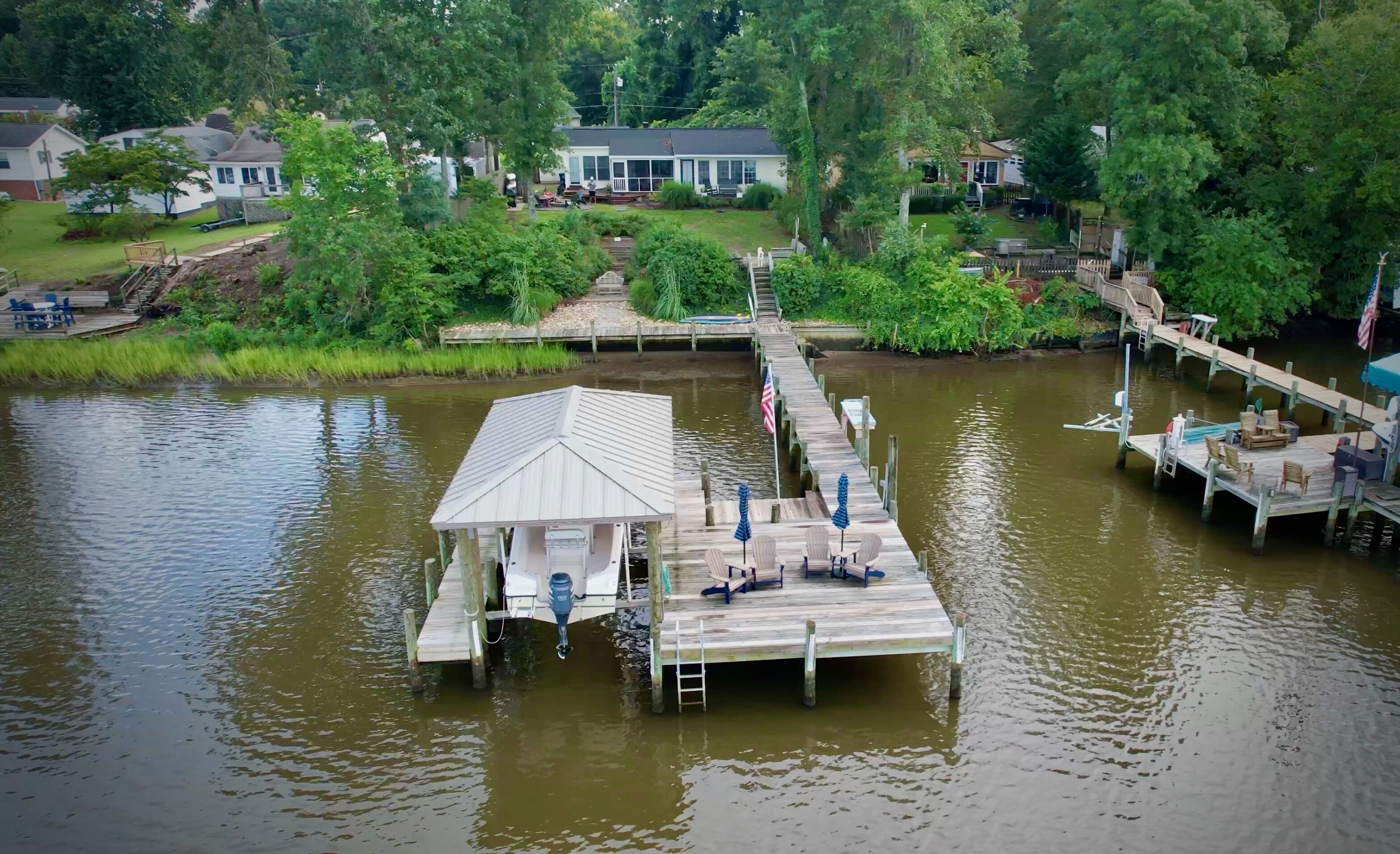 Een steiger die in het water uitloopt, met een overdekte constructie aan het einde, omgeven door begroeiing en woongebouwen.