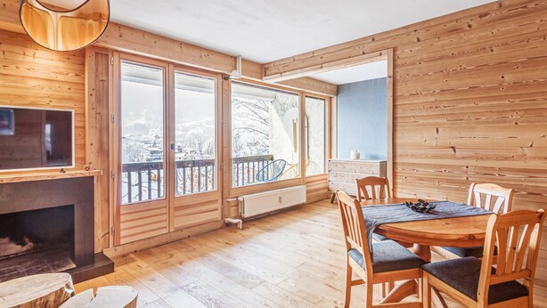 En ombonad träinredning med en öppen spis, matbord och stolar. Stora fönster ger utsikt över ett snötäckt landskap.