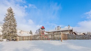 Ett snötäckt landskap med ett trästaket, en röd lekställning och en byggnad med ett sluttande tak.