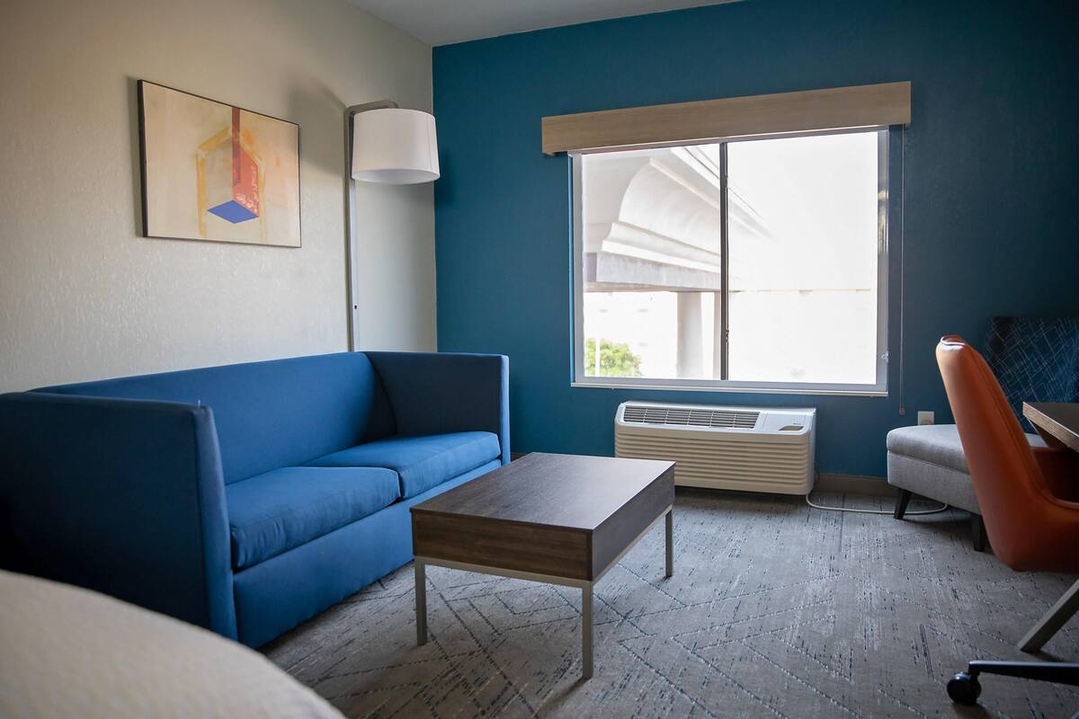 A room with a blue sofa, a wooden coffee table, a standing lamp, a window with blinds, and a radiator.