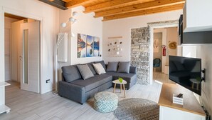 A modern living room with a wooden ceiling, grey sofa, wall art, and a stone wall. - GuestHost - Apartment of about 80 sqm, perfect for 4/6 people (but it can host up to 7 people), located in a pedestrian area, on the ground floor of a renovated house overlooking Lake Como. The property is located in the historic centre of the enchanting (Carate Urio)