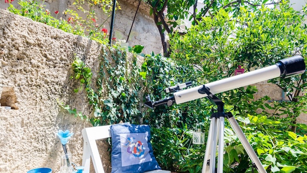 Un telescopio dispuesto sobre una silla blanca con cojín, un cubo azul y un pequeño farol sobre una mesa.