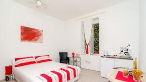 Habitación de hotel con cama, escritorio, silla, una mesita con aperitivos, un cuadro en la pared y una ventana con cortinas.