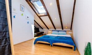 A bedroom with a sloped ceiling, wooden beams, a skylight, a bed with blue bedding, a black chair, and a small table.