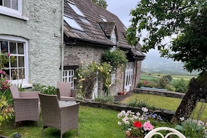 A stone house with a sloped roof, a garden with flowers, and a view of the countryside.