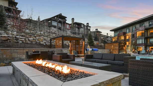 A stone fire pit with seating, a pool, and a stone wall with a wooden pergola.