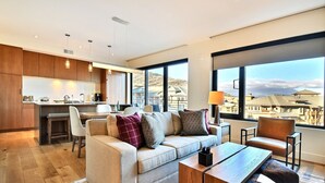 A modern living room with a sofa, coffee table, and dining area. There is a large window offering a view of buildings and hills.