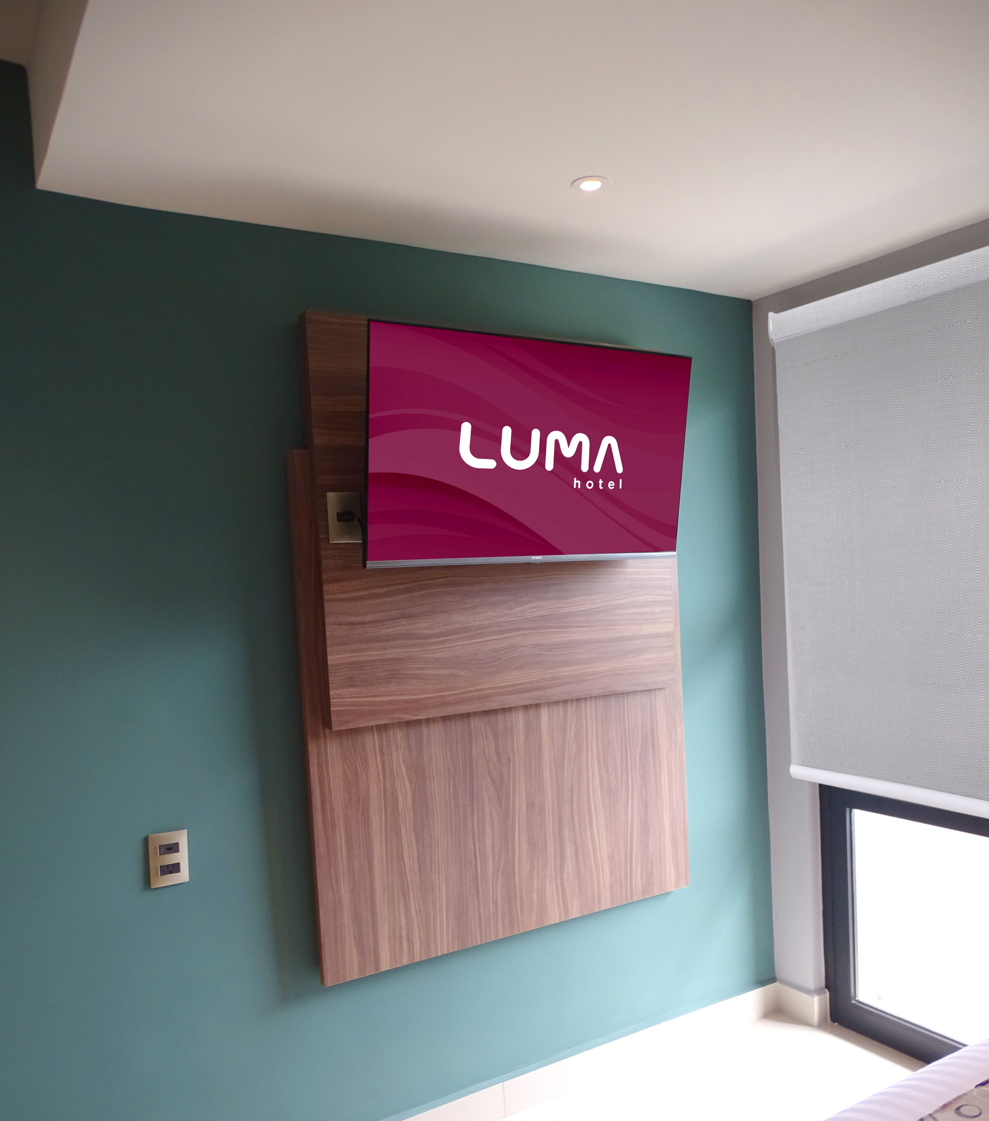 Televisão fixada na parede exibindo 'LUMA hotel' em uma parede verde-água com um painel de madeira abaixo.